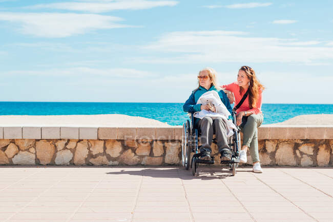 Cheerful adult daughter with elderly mother in wheelchair sitting stone fence along promenade near sea in summer looking away — Stock Photo