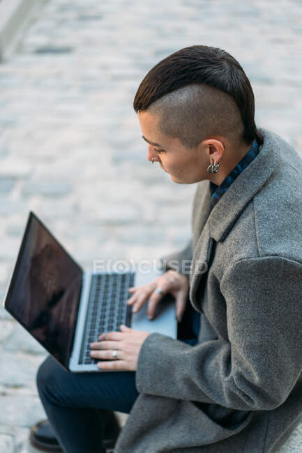 From above androgynous person with mohawk in boots and coat surfing internet on netbook while sitting in city — Stock Photo