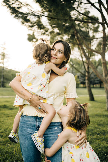 Усміхнена молода мати носить милу маленьку доньку в літньому одязі з браслетом і дивиться геть, стоячи на зеленому газоні в літньому парку — стокове фото