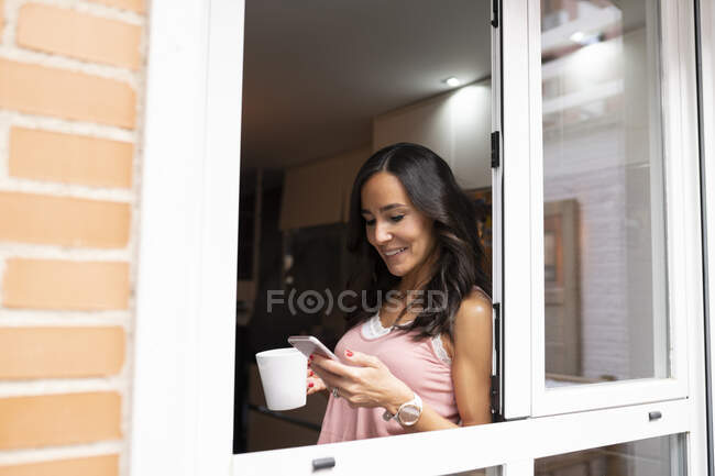 Красивая женщина, стоящая с чашкой утреннего кофе у окна и просматривающая мобильный телефон во время завтрака дома — стоковое фото