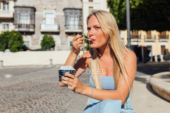 Красива блондинка молода жінка їсть холодне смачне морозиво, сидячи на вулиці міста в сонячний день влітку — стокове фото