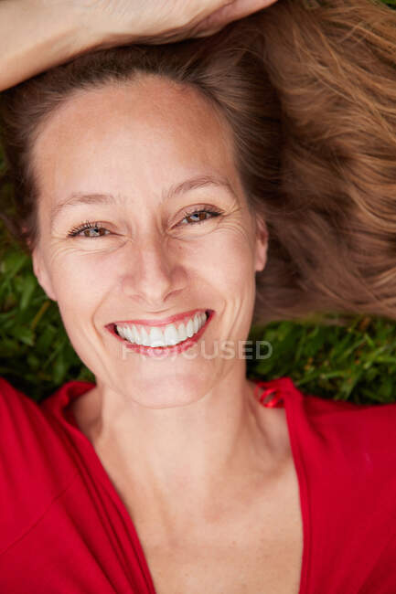 Femme vêtue de rouge couchée sur le sol dans un parc avec de l'herbe et regardant la caméra — Photo de stock