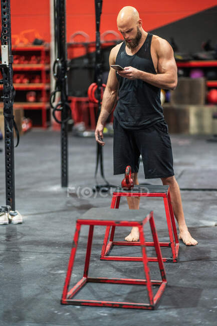 Musculoso deportista calvo de pie en el gimnasio moderno de navegación en el teléfono inteligente durante el descanso de entrenamiento - foto de stock