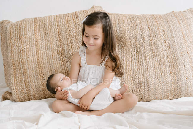 Petite fille joyeuse assise sur un lit doux à la maison et regardant bébé mignon — Photo de stock