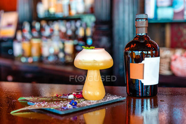 Sour cocktail with mint leaf in mushroom shaped glass served on wooden counter with bottle of alcohol drink in pub — Stock Photo