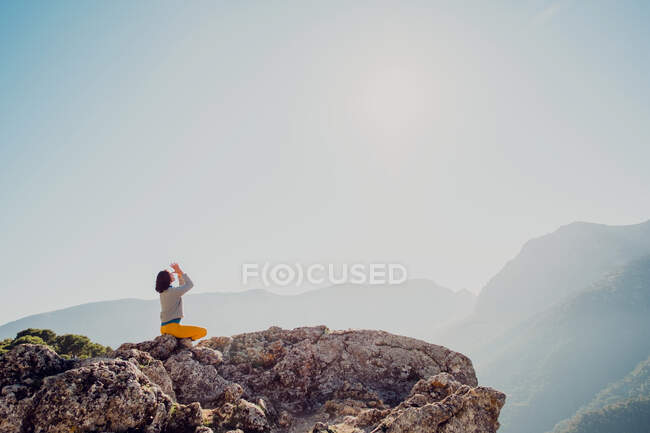 Вид сбоку анонимной женщины, сидящей на скале в горах и занимающейся йогой в Падмасане — стоковое фото