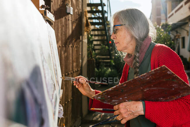 From below side view of aged female artists in protective mask painting on canvas in backyard on sunny day in summer — Stock Photo
