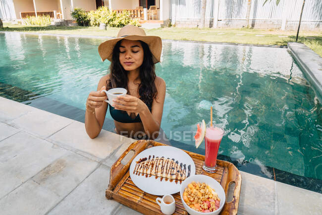 Touriste féminine joyeuse penchée au bord de la piscine tout en buvant du café contre plateau avec un délicieux petit déjeuner au soleil — Photo de stock