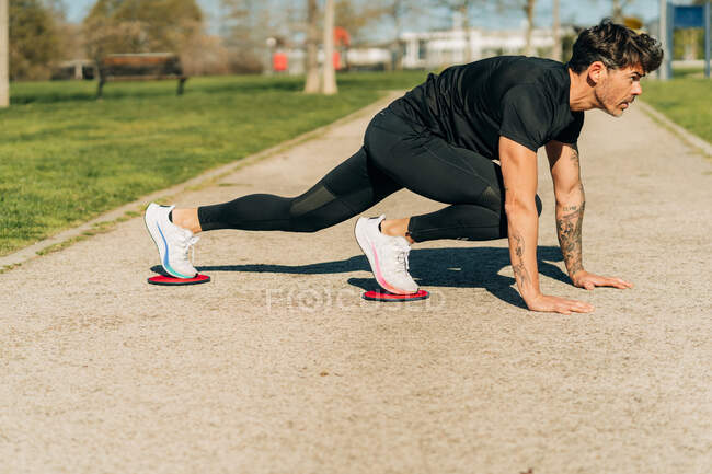 Nivel del suelo de atleta masculino atento en pose de tabla con entrenamiento de discos deslizantes y mirando hacia adelante en el pavimento en la ciudad - foto de stock