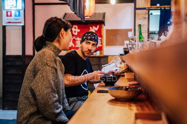 Vista lateral de la mujer y el hombre comunicándose mientras come comida asiática en el mostrador de madera en la cafetería moderna - foto de stock