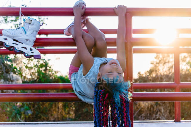 Fröhliches ethnisches Kind mit hellen Zöpfen, die mit Rollschuhen am Zaun hängen und im Sonnenlicht wegschauen — Stockfoto