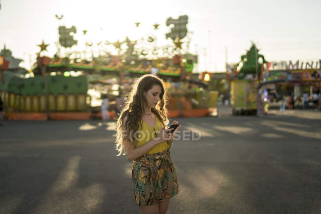 Mujer con el pelo ondulado de pie en el recinto ferial y el teléfono móvil de navegación al atardecer en verano - foto de stock