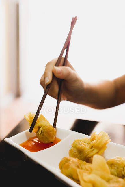 Рука невпізнаваної жінки, використовуючи палички для занурення смачного вонтона в соус на розмитому фоні ресторану — стокове фото