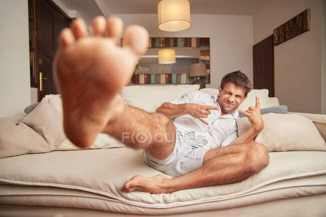 Повне тіло позитивного молодого босоніж чоловіка в повсякденному одязі, що досягає ніг до камери і жестикулює знак рогів, розслабляючись на зручному дивані вдома — стокове фото