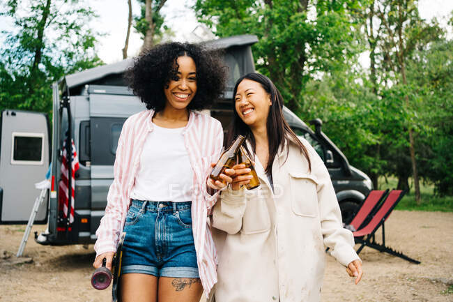 Happy young multiethnic female friends clinking bottles of beer while having fun and enjoying summer holidays together near camper van parked in nature — Stock Photo