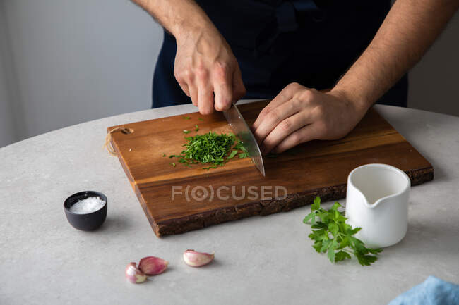 Unrecognizable male in apron cutting fresh parsley on wooden cutting board near salt and garlic while cooking lunch at home — Stock Photo
