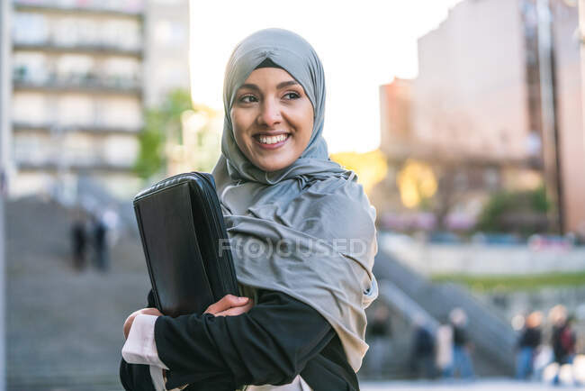 Empreendedora muçulmana alegre no hijab e com pasta de pé olhando para longe na rua — Fotografia de Stock