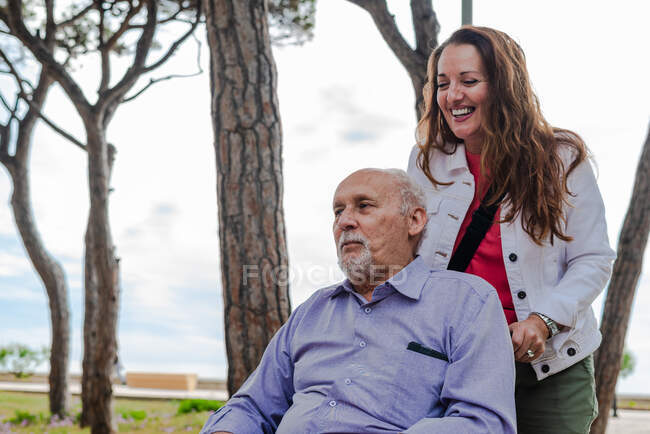 Позитивная взрослая дочь толкает инвалидное кресло с престарелым отцом во время прогулки в парке летом — стоковое фото