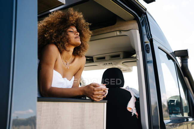 Low angle of relaxed African American female with mug of hot drink closing eyes and chilling inside modern RV in summer morning — Stock Photo