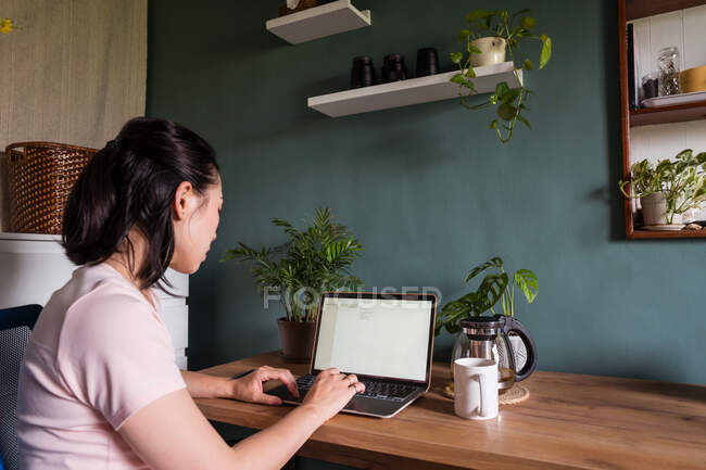 Вид сбоку азиатской женщины-фрилансера, читающей документы на ноутбуке, сидя за столом во время удаленной работы — стоковое фото