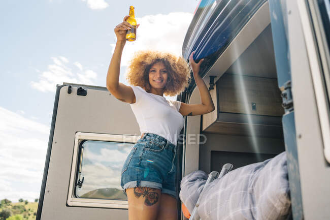 Vista lateral de la mujer negra encantada con la cerveza riendo y mirando a la cámara mientras está de pie en la escalera fuera de autocaravana y subiendo a la cama en el día de verano en el campo - foto de stock