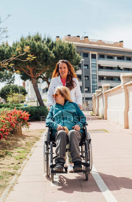 Happy adult woman looking at camera while pushing wheelchair with senior mother during walk on the street in city in summer — Stock Photo