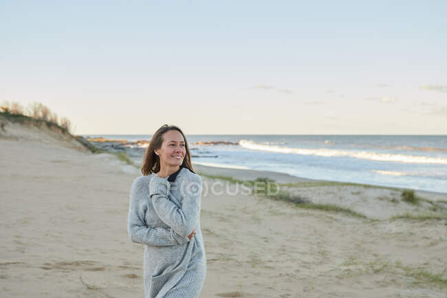 Вміст жінки в теплому одязі, що стоїть на пляжі біля моря і насолоджується літнім вечором, дивлячись далеко — стокове фото