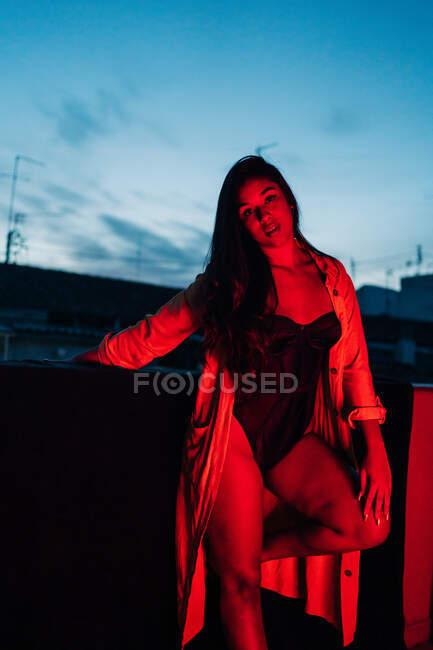 Приємна молода іспанська етнічна жінка в білизні дивиться на камеру, відпочиваючи на терасі під червоним неоновим світлом вночі. — стокове фото