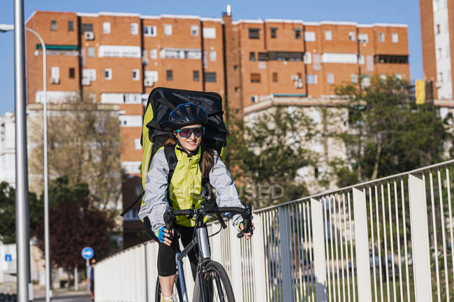 Веселая курьерша с термальной сумкой на велосипеде по мосту во время доставки еды в город — стоковое фото
