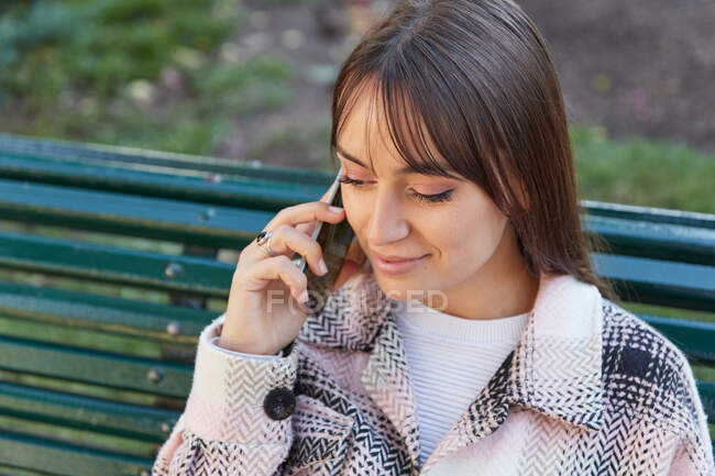 От современной женщины тысячелетия в стильном весеннем наряде, сидящей на скамейке и отвечающей на телефонный звонок, отдыхающей на городской улице и смотрящей в сторону — стоковое фото