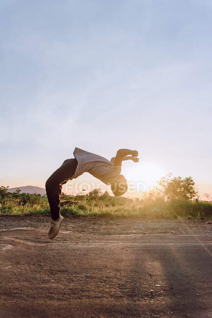 Vista lateral del macho acrobático saltando por encima del suelo y realizando peligrosos trucos de parkour en un día soleado - foto de stock