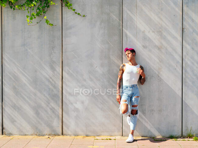 Trendy gay maschio con rosa capelli e strappato jeans appoggiato su muro in città e guardando lontano — Foto stock