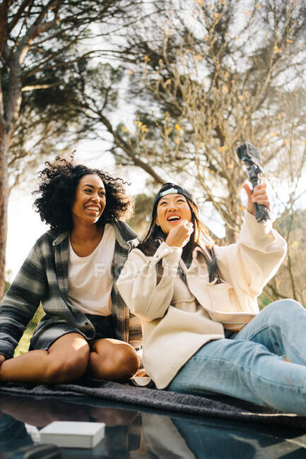 Baixo ângulo de jovens namoradas multirraciais alegres sentadas no telhado da van campista e tirando fotos ao passar férias de verão juntos no campo — Fotografia de Stock