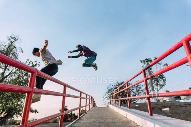 Low angle side view of fearless male friends jumping above metal railing in city while performing parkour stunt on sunny day — Stock Photo