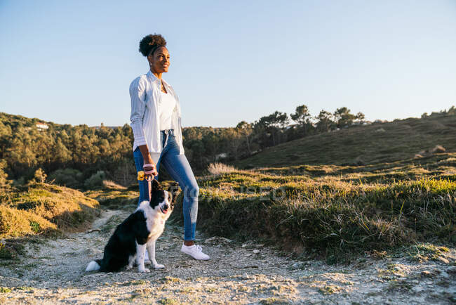 Vista lateral de cuerpo completo de una mujer étnica feliz con perro Border Collie caminando juntos por el sendero entre colinas cubiertas de hierba en la soleada noche de primavera - foto de stock