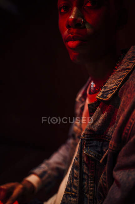 Crop calm stylish African American man in jeans jacket under neon red light in shade on black background looking at camera — Stock Photo