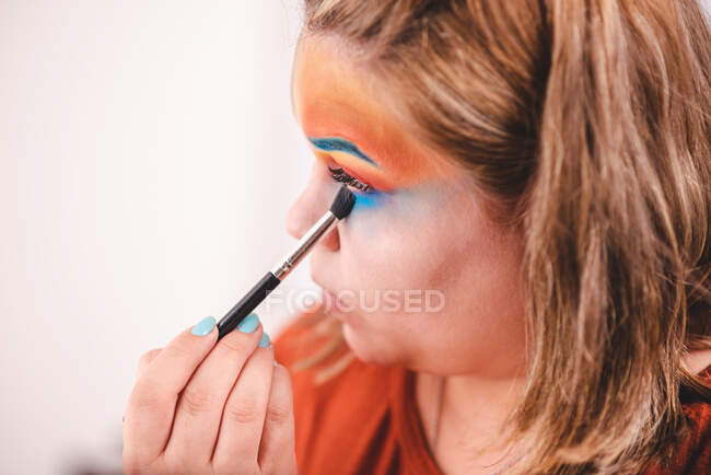 Overweight female with palette applying colorful pigments on face while looking at mirror near ring light in studio — Stock Photo