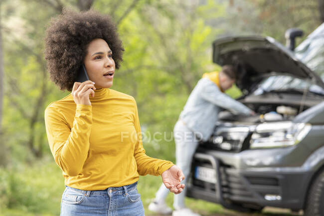 Mulher negra chocada fazendo chamada para o serviço de emergência, enquanto jovem tentando consertar o motor da van durante a viagem de carro no campo — Fotografia de Stock