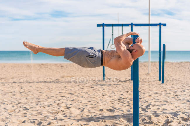 Vista lateral do homem sem camisa focado pendurado em barras paralelas segurando seu peso em linha reta horizontal contra a praia à luz do sol — Fotografia de Stock