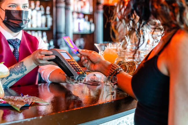 Female bartender with POS terminal standing at counter with crop anonymous woman paying with plastic card for order in bar — Stock Photo