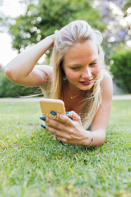 Sonriente encantadora hembra acostada en el césped en el parque y escuchando música en los auriculares en verano - foto de stock