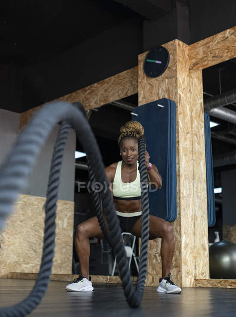Strong African American female athlete in activewear exercising with battle ropes while looking forward during high intensity training in gym — Stock Photo