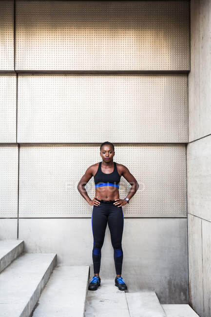 Femme afro-américaine positive en vêtements de sport en gardant les mains sur la taille et en regardant la caméra tout en se tenant debout sur les escaliers pendant l'entraînement extérieur — Photo de stock