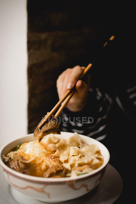 Mão com pauzinhos puxando macarrão enquanto come sopa de macarrão de carne de grande tigela de cerâmica — Fotografia de Stock