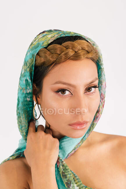 Modelo femenino joven con peinado trenzado y maquillaje elegante con pañuelo colorido con estampado floral verde mirando a la cámara sobre fondo blanco - foto de stock