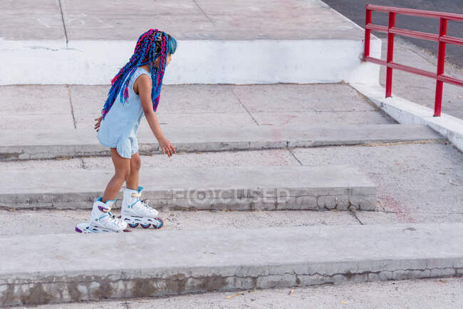 Vista lateral da criança étnica em patins com tranças coloridas em pé na escada — Fotografia de Stock