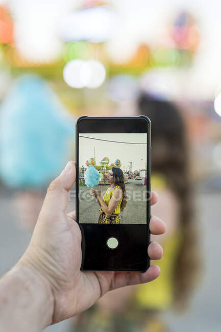 Cortar pessoa anônima usando smartphone e tirar foto de fêmea com algodão doce azul se divertindo no parque de diversões — Fotografia de Stock
