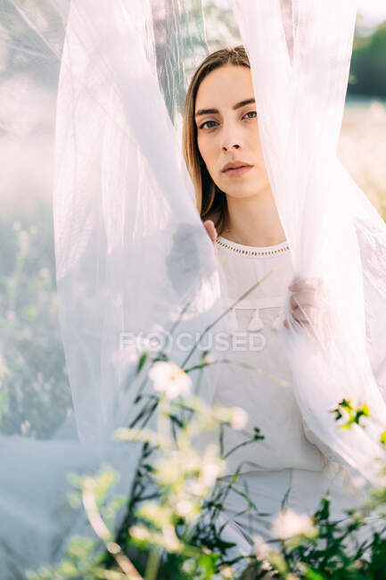 Gentle young female in white dress sitting in white light tulle with flowers looking at camera on countryside field — Stock Photo