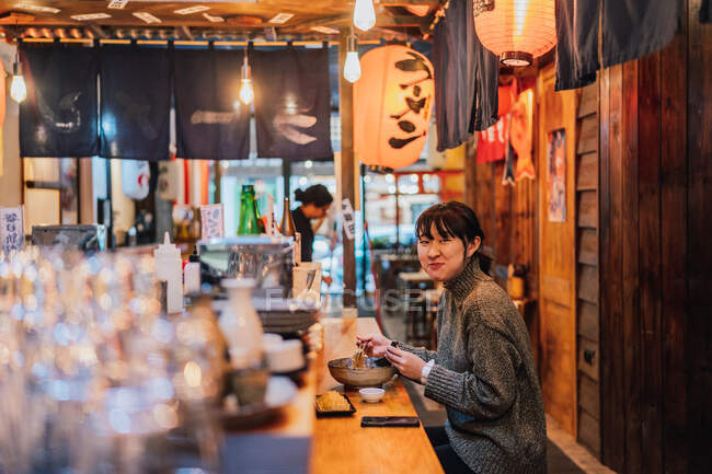 Vista lateral de la mujer asiática satisfecha en suéter sonriendo mientras toma cuchara de trabajador sentado en el mostrador de madera en el bar de ramen - foto de stock