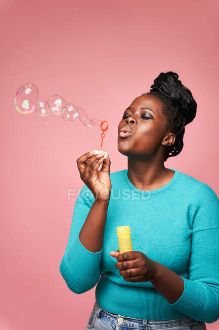Happy African American female looking down wearing blue clothes and blowing soap bubbles against pink background in studio — Stock Photo